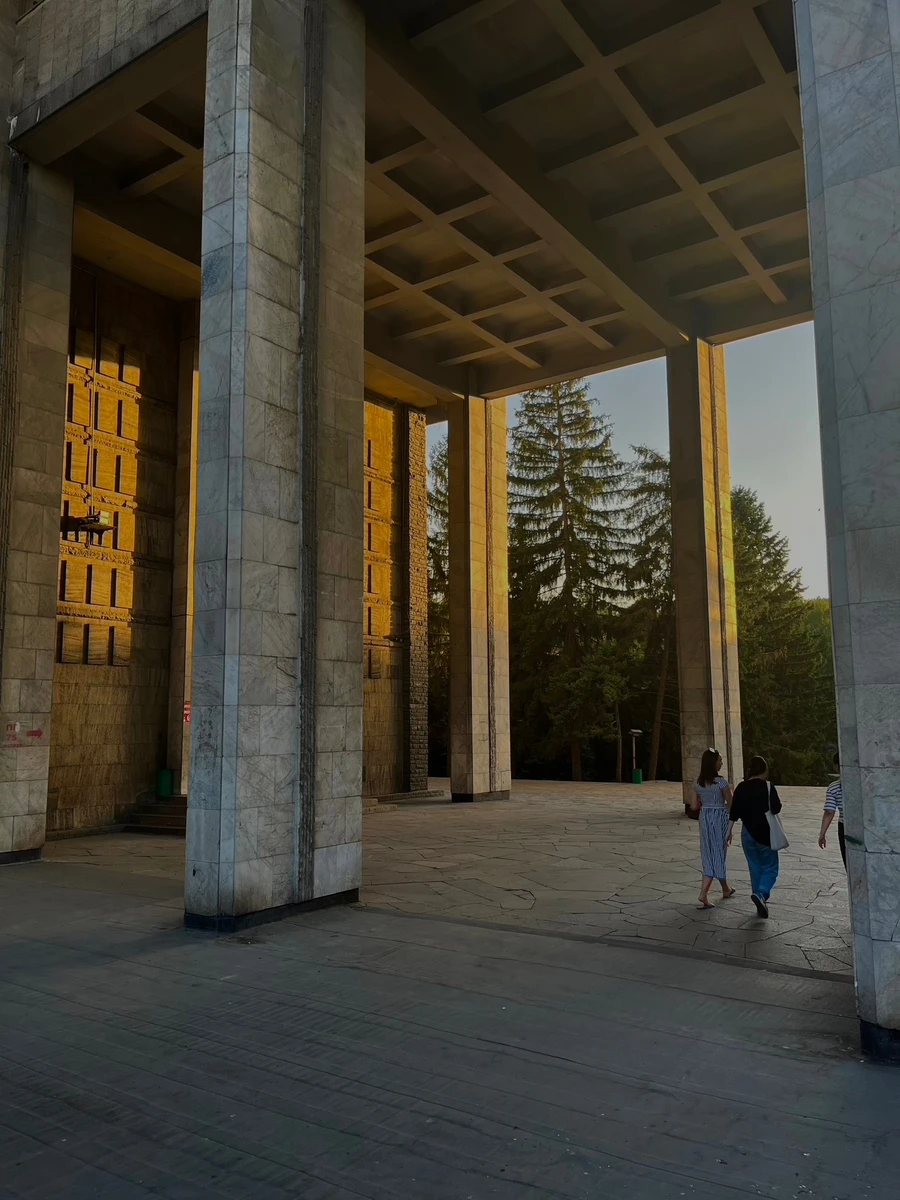 People walking beneath the massive stone colonnade of an Almaty monument at golden hour